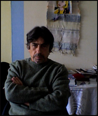 Portrait of director José Vieira. He appears to be sitting with his arms crossed. The photo only captures him from the torso up. He is in a room with white walls and blue details. Behind him there is an out of focus table with objects and documents.